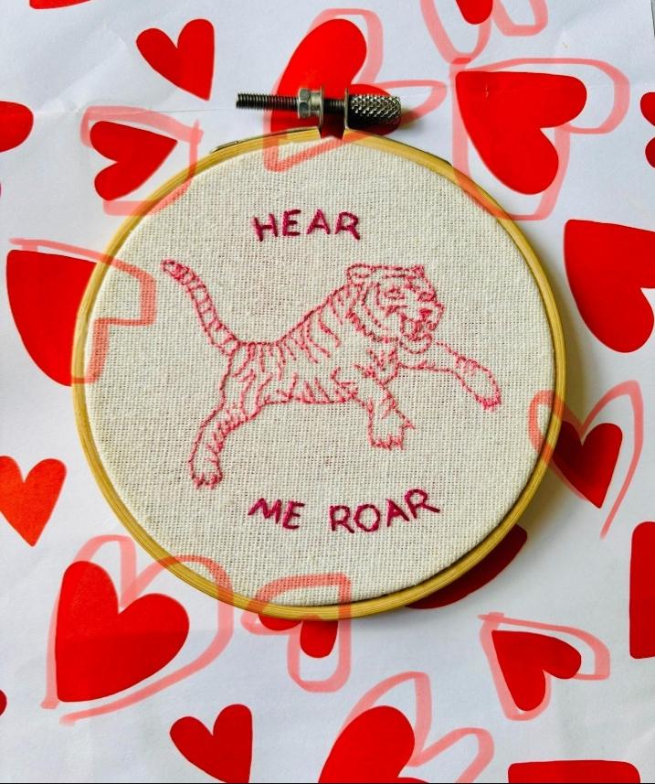 Geborduurde springende tijger en tekst, op stof in een borduurring. Daaromheen hartjes. Hoofdkleur rood.